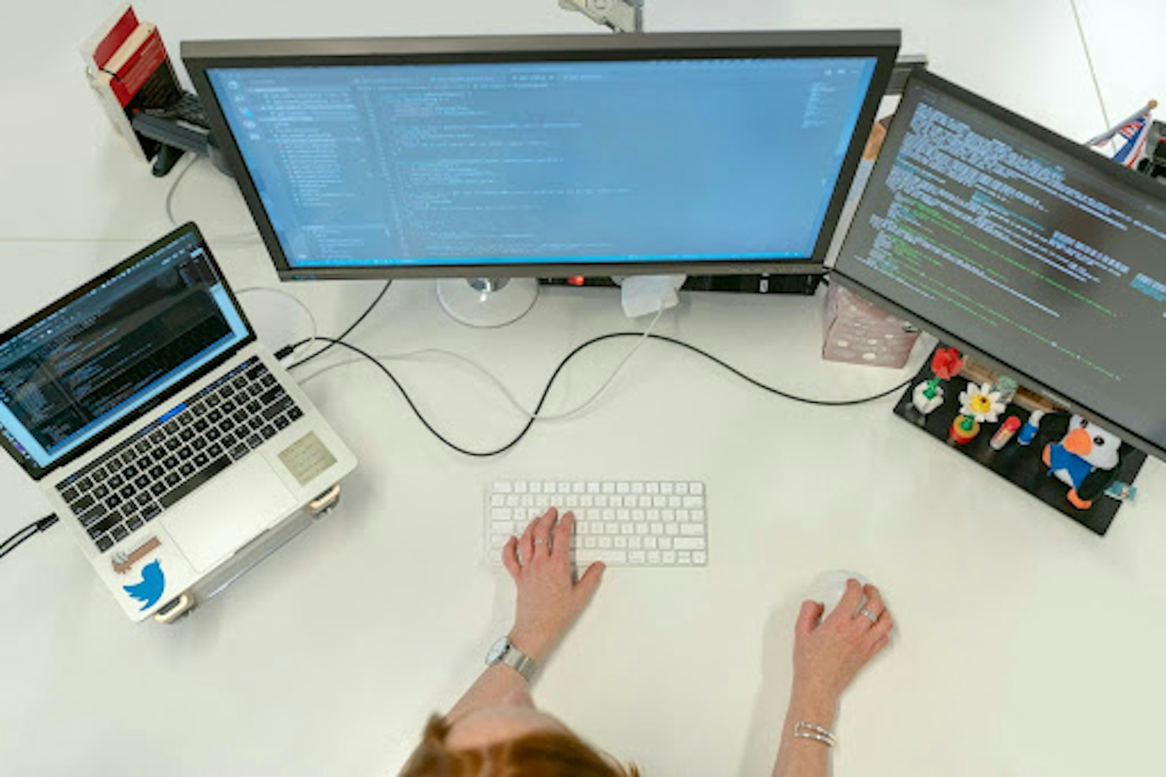 Aerial view of a computer desk with three large screens, one small keyboard and two hands controlling them.