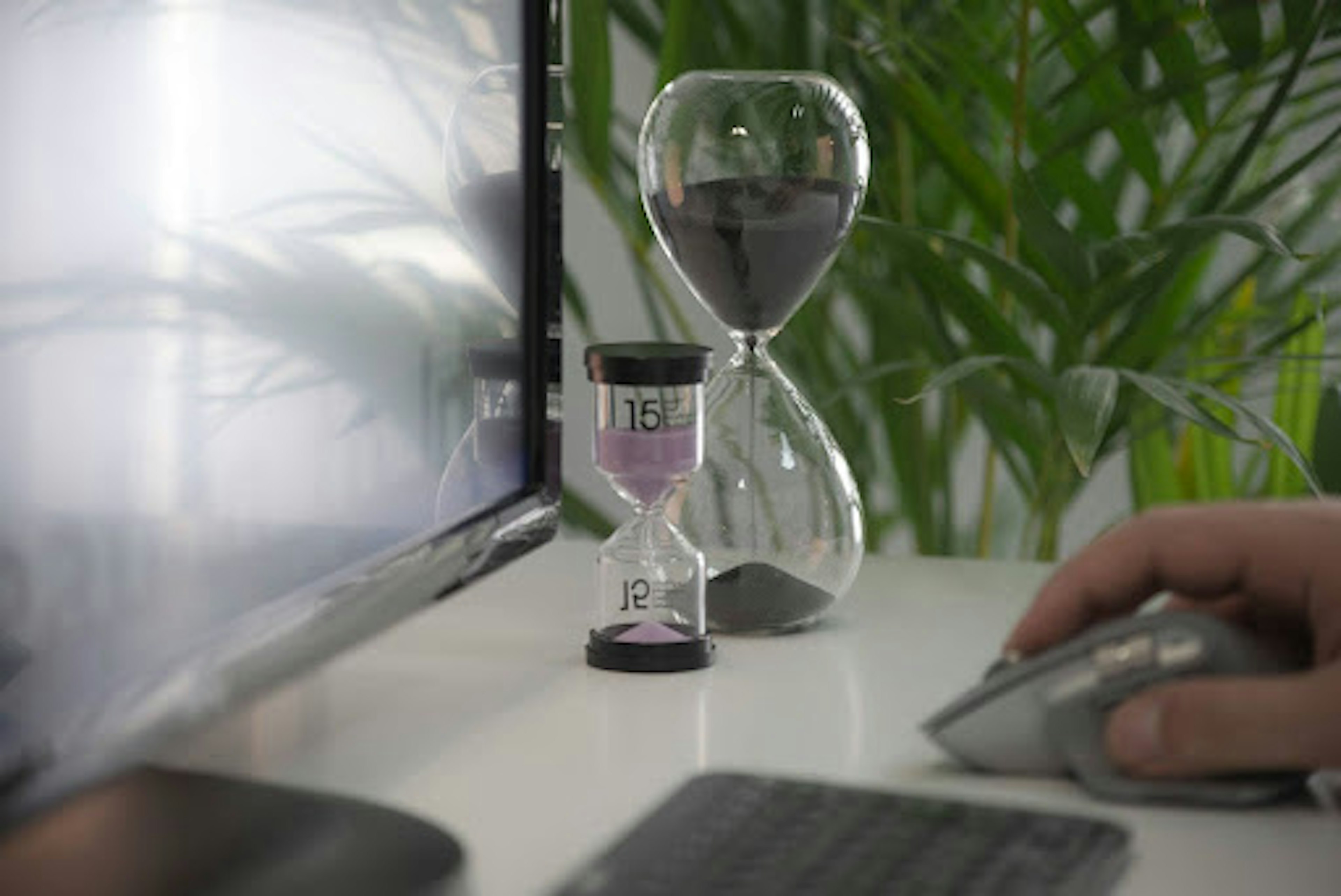 Two hourglasses next to a computer screen. The foreground has a hand on a mouse and a keyboard.