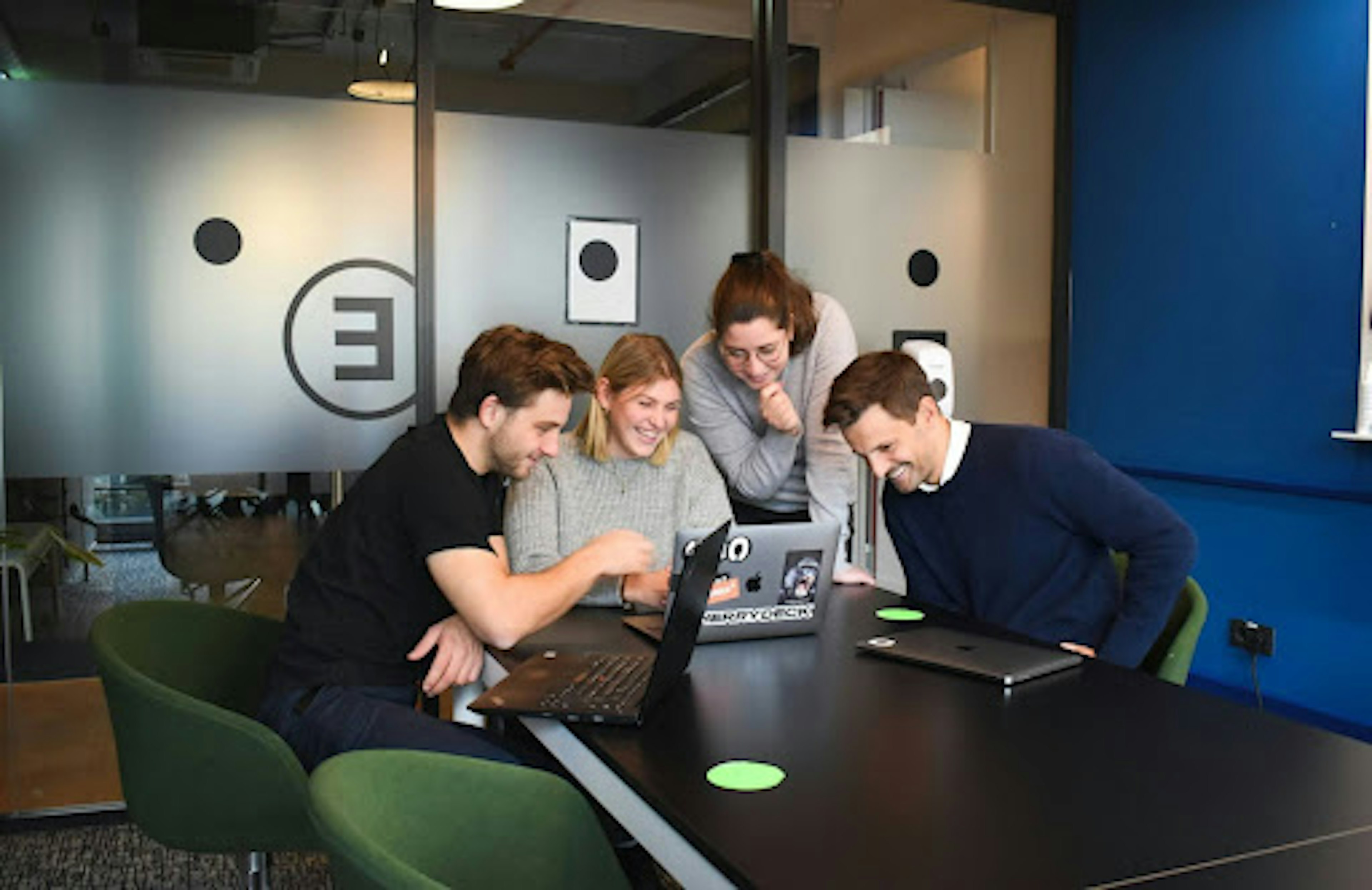 Two women and two men in a conference room gathered around a laptop and smiling.